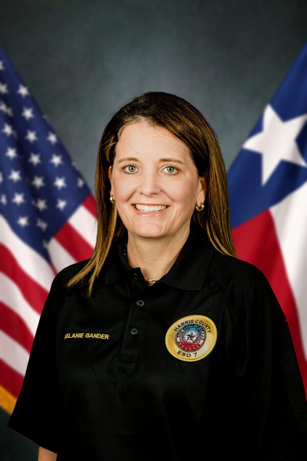 Melanie Gander in a black polo shirt with a Harris County ESD 7 badge, standing in front of the US and Texas flags.