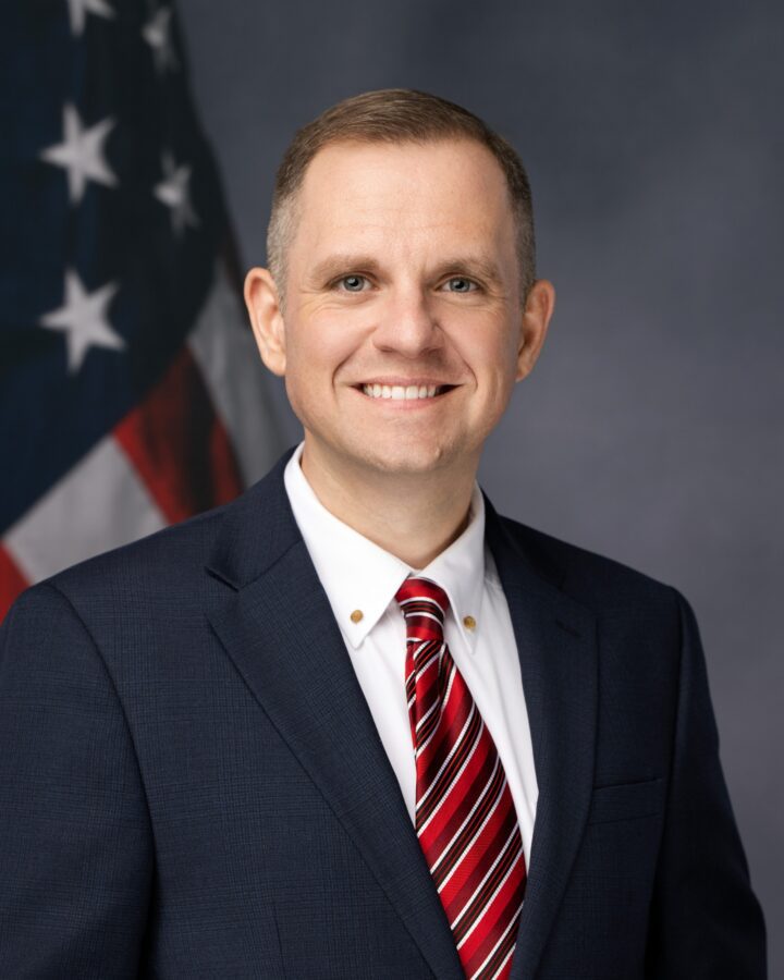 A man in a navy suit, white shirt, and red striped tie stands in front of a U.S. flag, smiling at the camera against a neutral background.