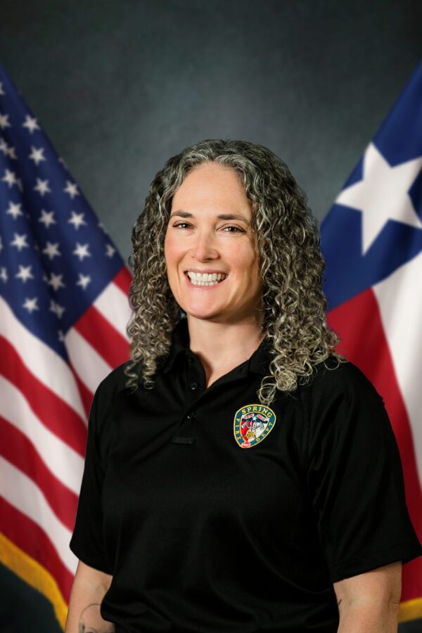 A person with curly gray hair smiles in front of U.S. and Texas flags, wearing a black shirt with an official patch.