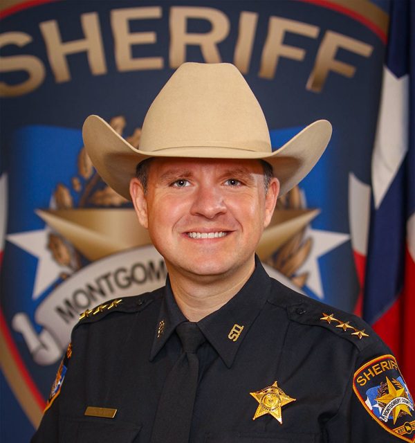 A sheriff in uniform and a cowboy hat smiles in front of a backdrop displaying the sheriff’s badge and insignia.