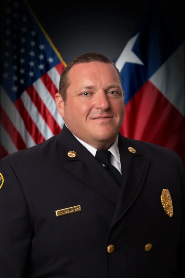 A man in a formal fire department uniform poses in front of the American and Texas flags.