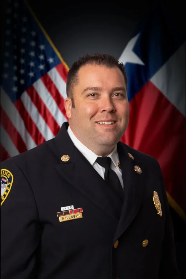 A man in a formal fire department uniform stands in front of the American and Texas flags, smiling at the camera.