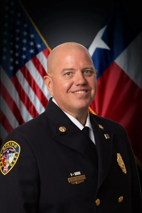 A man in a formal fire department uniform poses in front of the American and Texas state flags.
