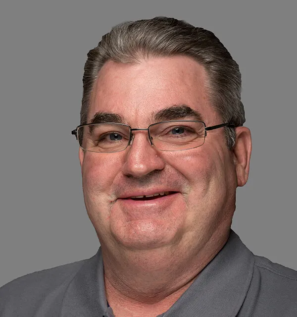 Smiling middle-aged man with short gray hair, glasses, and a gray collared shirt against a plain gray background.
