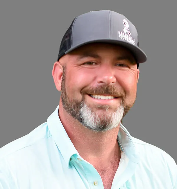 A man with a beard and mustache wearing a black baseball cap and a light blue shirt, smiling at the camera against a plain gray background.
