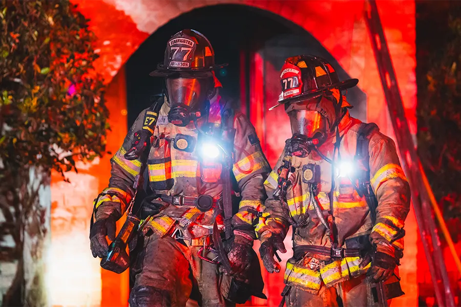 Two firefighters in full gear and helmets walk side by side at night, illuminated by red emergency lights, with a building entrance in the background.