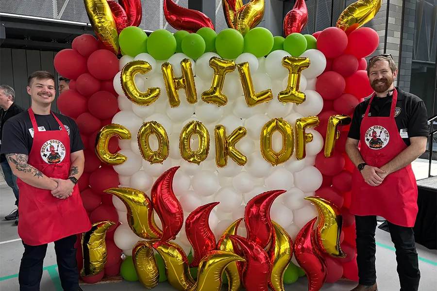 Two men in red aprons stand in front of a festive balloon display reading "Chili Cook Off" with balloon flames and red, white, and green balloons.