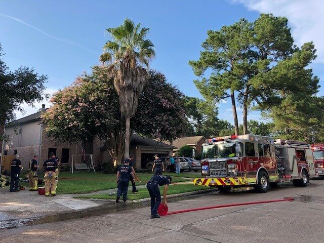 Firefighters work outside a residential house, with a fire truck parked on the street and hoses laid out on the ground.