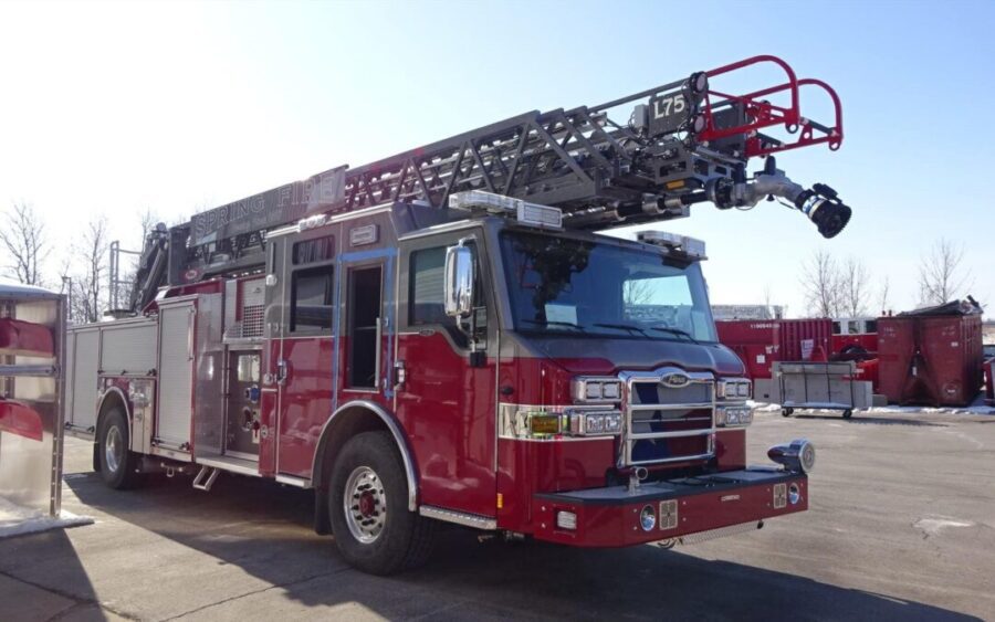Red fire truck with an extended aerial ladder parked on a paved surface, with industrial buildings and leafless trees in the background.