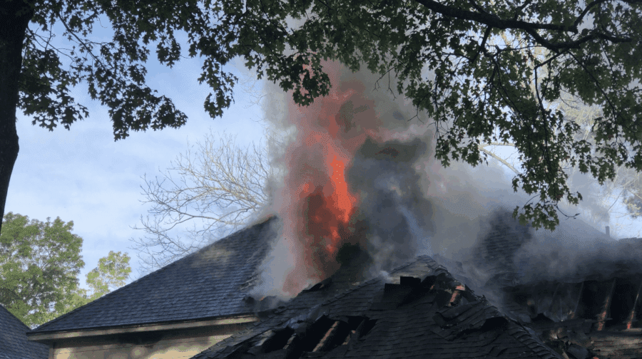 A house roof is on fire with bright flames and thick smoke rising through a hole, surrounded by trees and a clear sky.