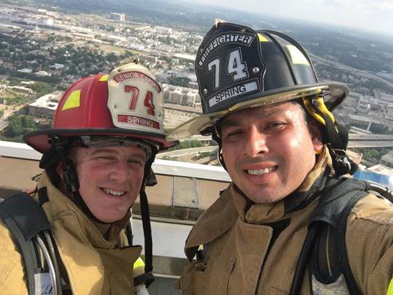 Two firefighters in uniform stand on a rooftop with a cityscape in the background, both wearing helmets labeled "74 Spring.
