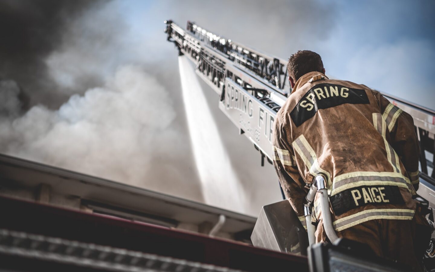 A firefighter in full gear operates a fire hose from a ladder truck, spraying water toward a smoky building.
