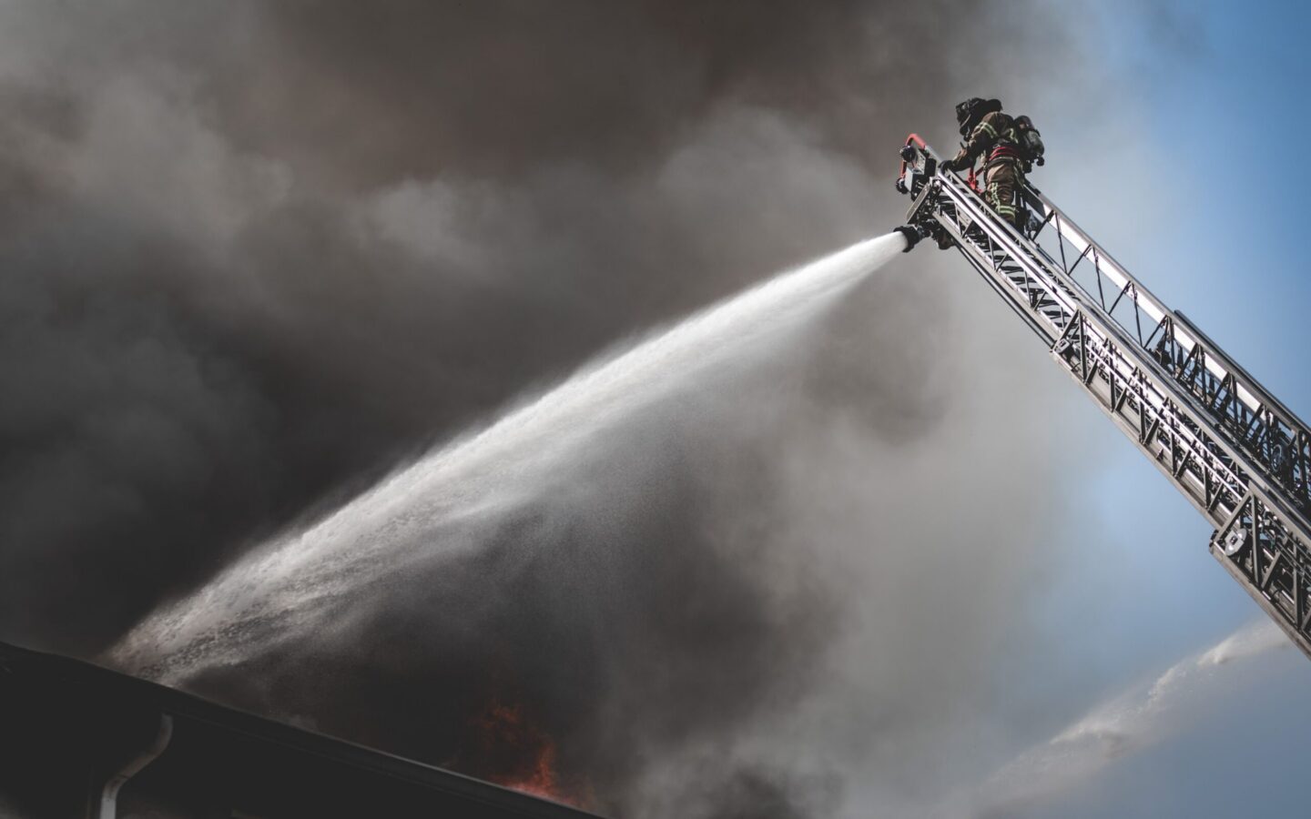 A firefighter on an extended ladder sprays water onto a fire below, with thick smoke filling the sky in the background.