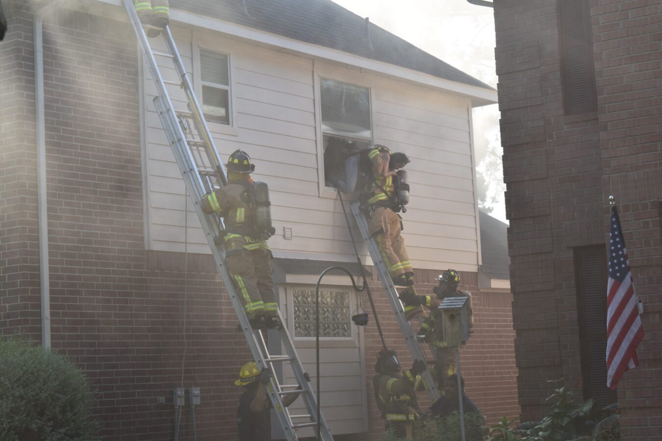 Firefighters in full gear use ladders to access the second-floor window of a smoke-filled house, with an American flag visible nearby.
