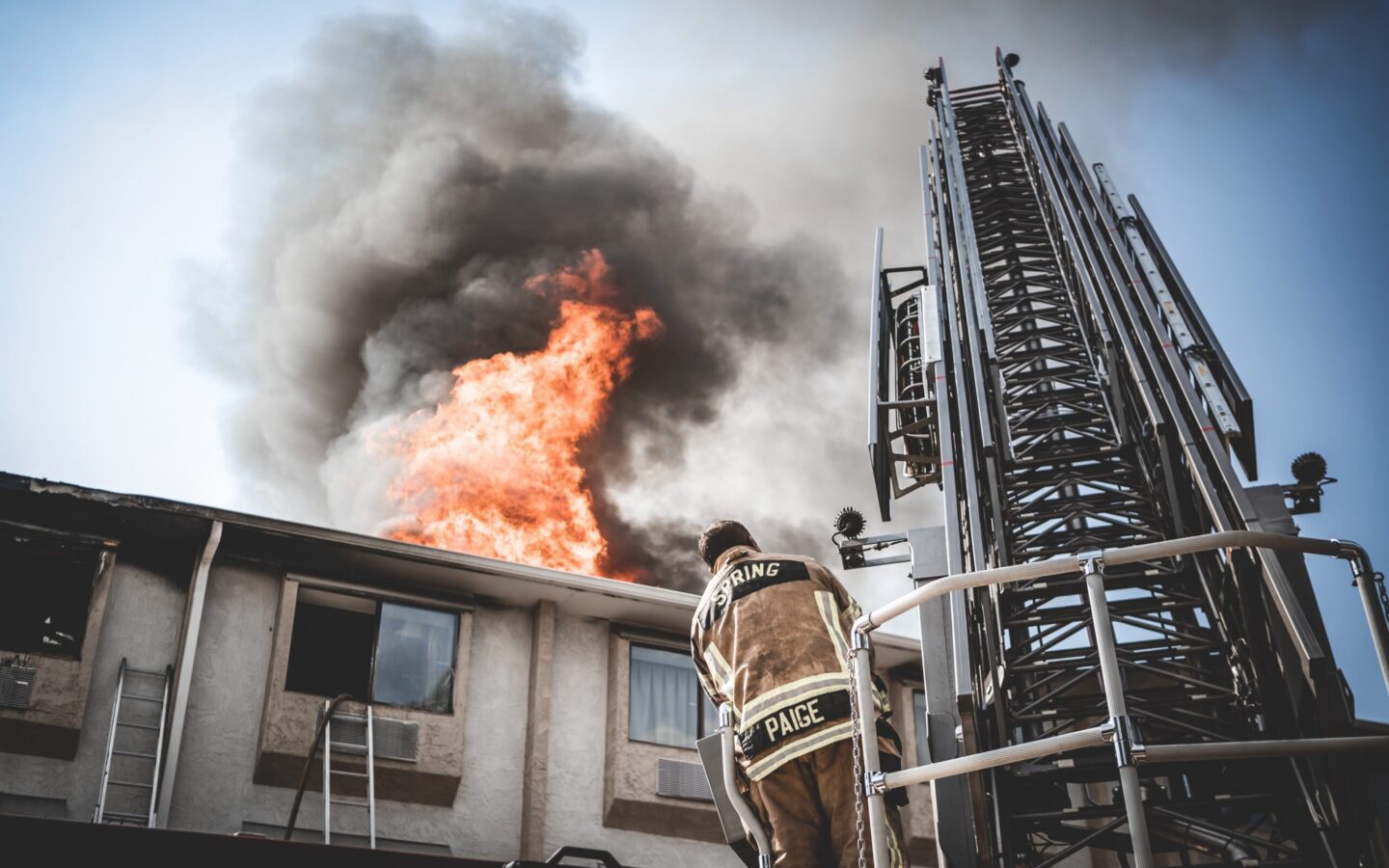 A firefighter stands on a ladder truck platform facing a burning building with flames and thick smoke coming from the roof.