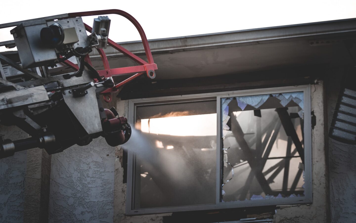 A fire hose on a ladder sprays water through a broken window of a building, with visible fire damage and shattered glass.