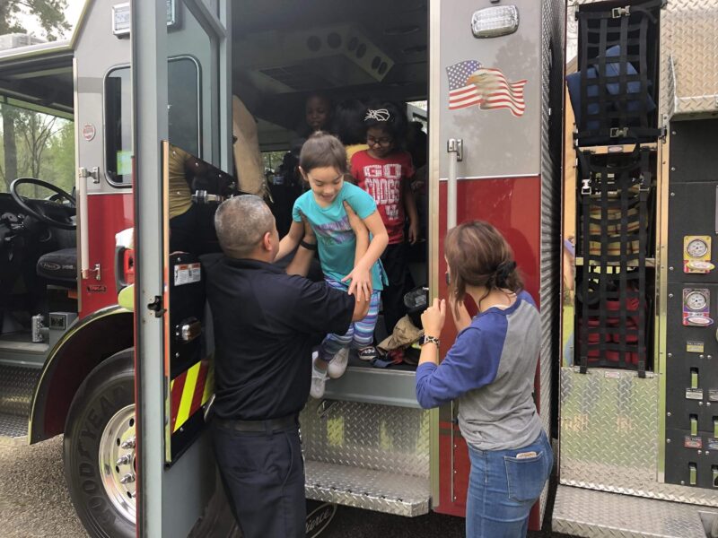 A firefighter helps a young girl step out of a fire truck while a woman stands nearby; other children are visible inside the truck.