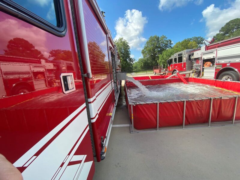 A fire truck pumps water into a large portable tank, with other fire trucks visible in the background on a sunny day.