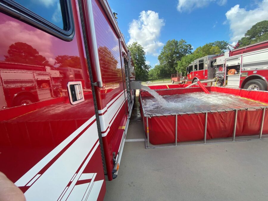 A fire truck pumps water into a large portable tank, with other fire trucks visible in the background on a sunny day.