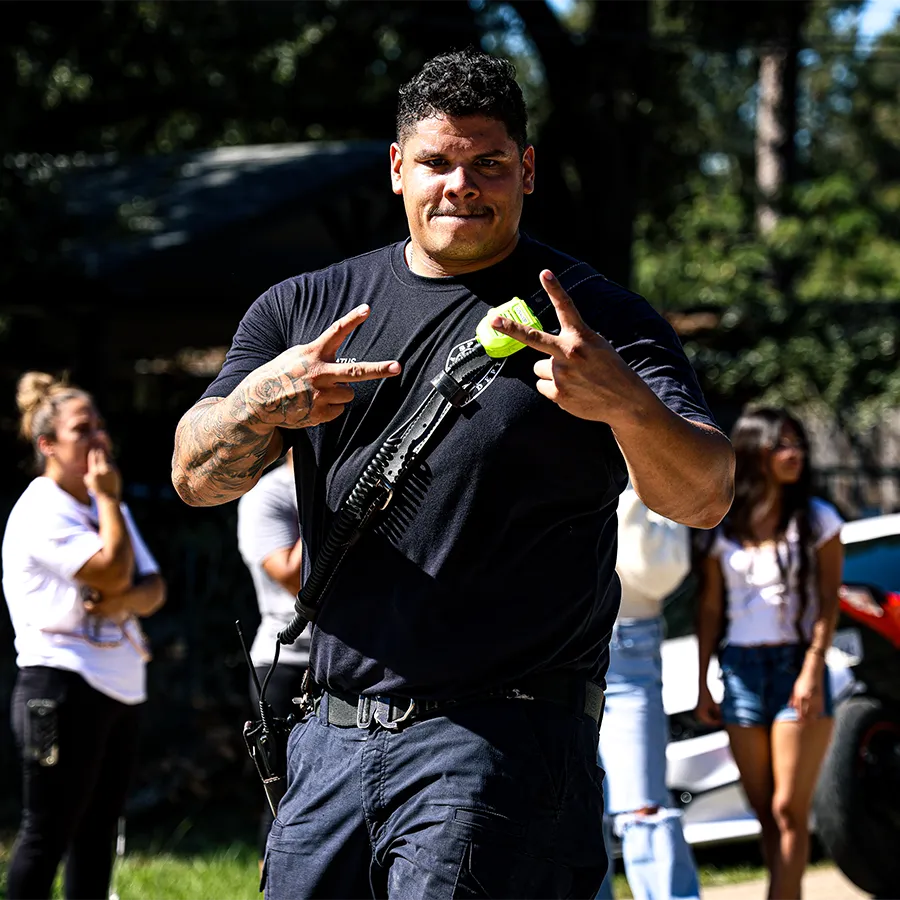 A man in a dark shirt and cargo pants, carrying a radio, flashes peace signs with both hands outdoors. Several people and parked cars are visible in the background.