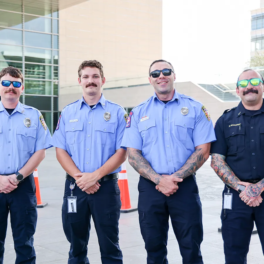 Four uniformed firefighters with mustaches stand side by side outside a modern building, smiling at the camera with their hands clasped in front.