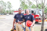 Two firefighters in uniform stand in front of a fire truck, one holding a water bottle and the other holding a helmet, in a parking lot with trees in the background.