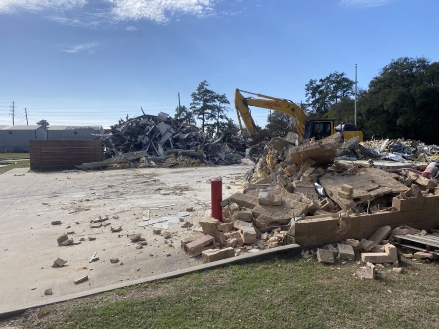 A yellow excavator moves debris at a demolition site with piles of rubble, broken bricks, and concrete under a clear blue sky.
