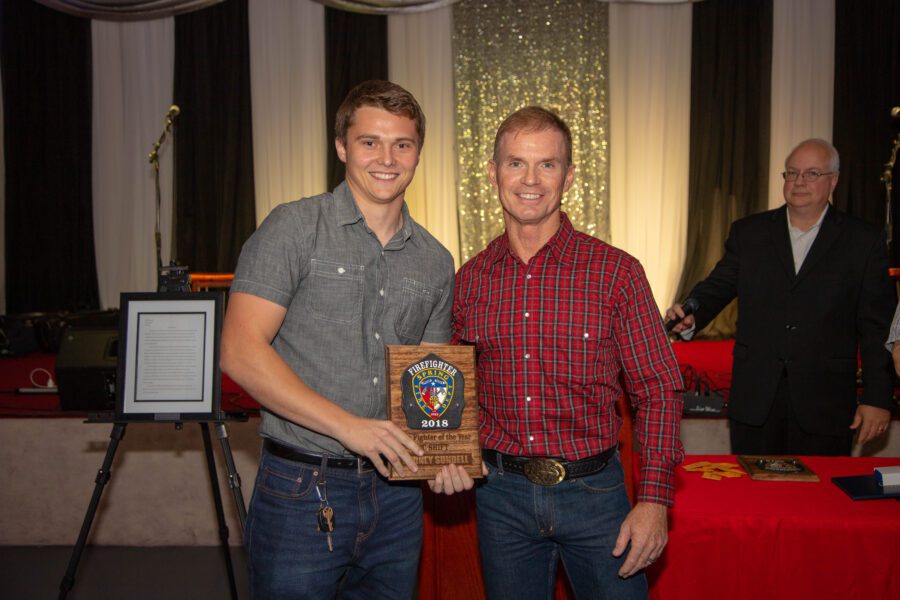 Two men stand indoors, one holding a 2018 award plaque. Both are smiling at the camera. A framed document and another man are visible in the background.