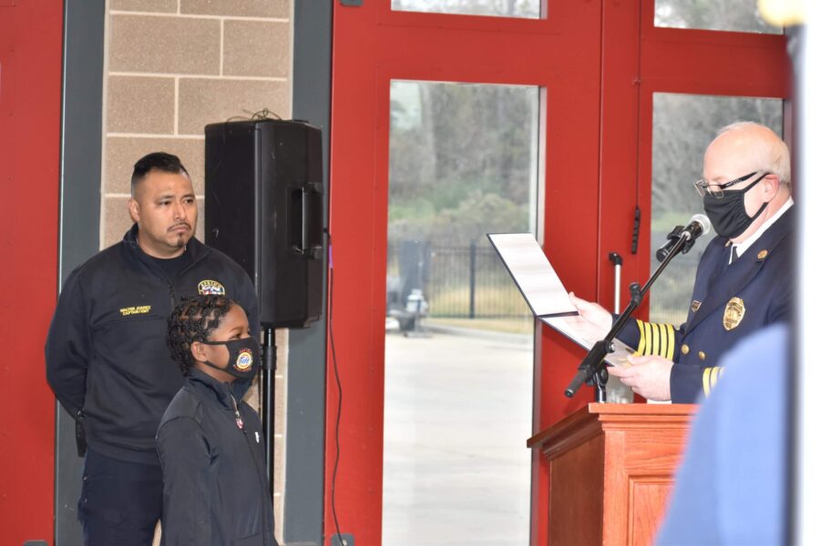A uniformed official reads from a document at a podium while a man and a child, both in dark uniforms, stand nearby inside a building.