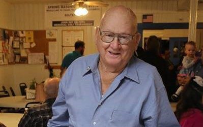 An older man wearing glasses and a light blue shirt smiles while standing in a community room with several people in the background.