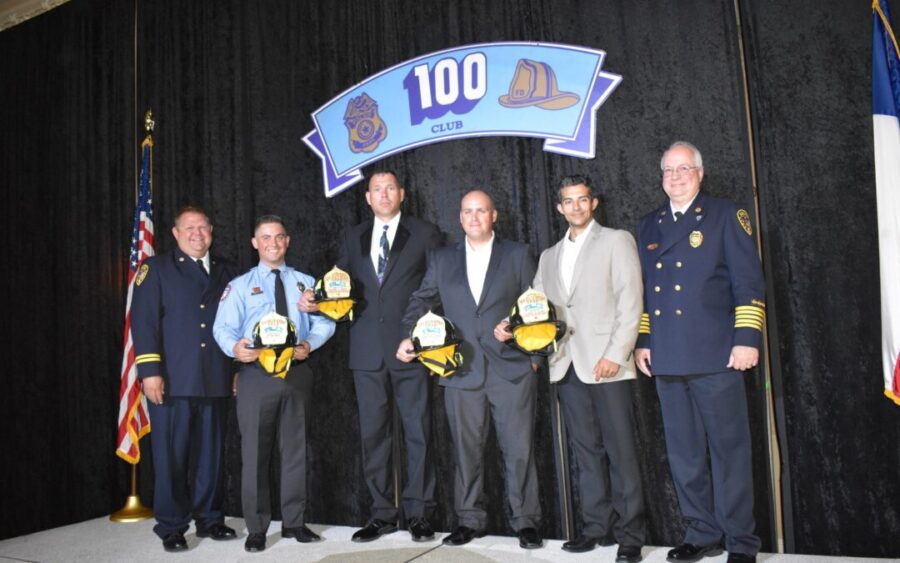 Six men stand on stage at a 100 Club event, four holding firefighter helmets, flanked by two men in uniform, with an American flag and a banner in the background.
