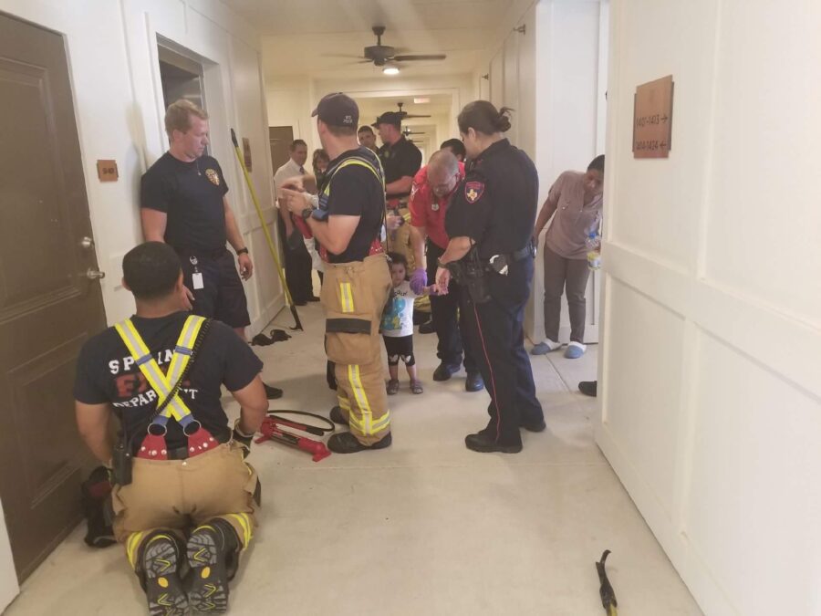 Firefighters and police officers interact with children and adults in a hallway, with rescue tools on the floor and people watching in the background.