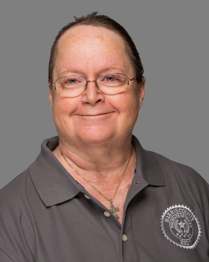A person with short hair and glasses wears a gray Harris County Emergency Services District polo shirt, smiling at the camera against a plain gray background.