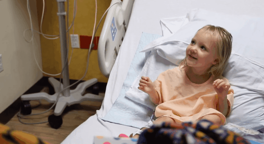 A young child sits upright in a hospital bed, wearing a hospital gown and smiling, with medical equipment visible in the background.