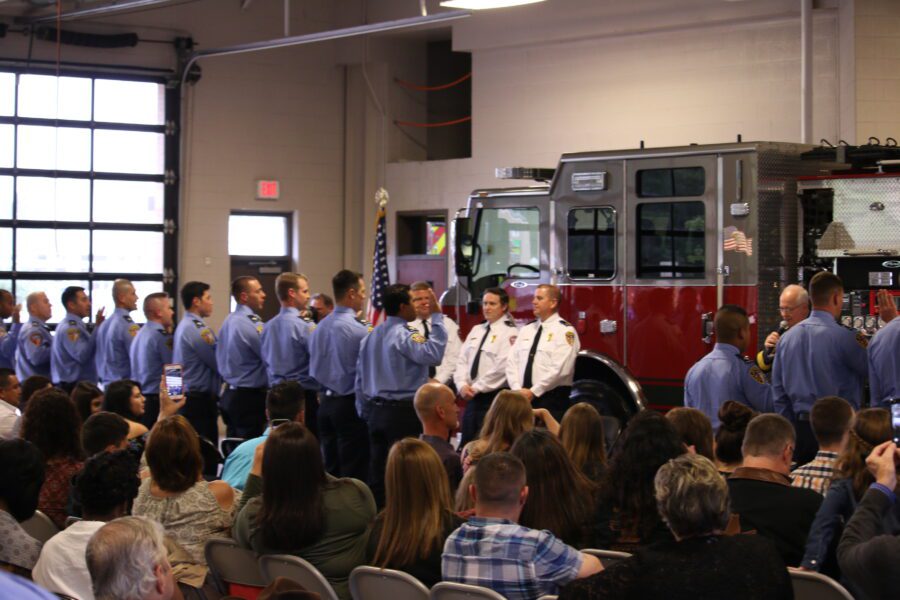 A group of firefighters in uniform stand in front of a fire truck during a ceremony, with an audience seated and watching.