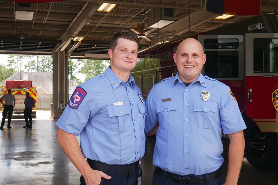 Two firefighters in uniform stand and smile inside a fire station, with fire trucks and other personnel visible in the background.