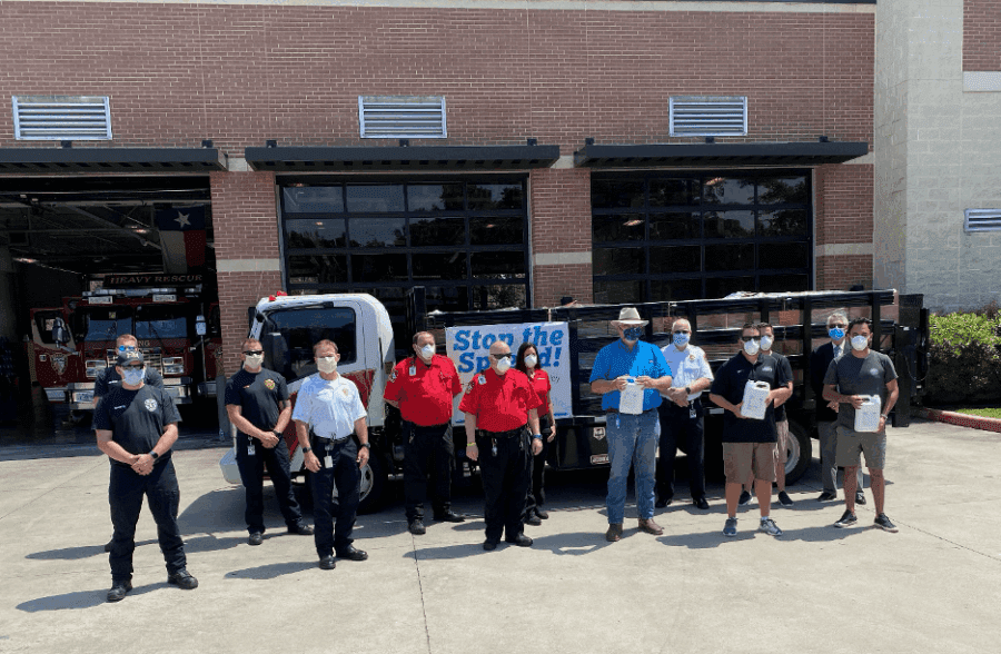 A group of people wearing masks stands in front of a fire station and a truck with a "Stop the Spread!" sign. Some hold papers or boxes. Fire trucks are visible in the background.