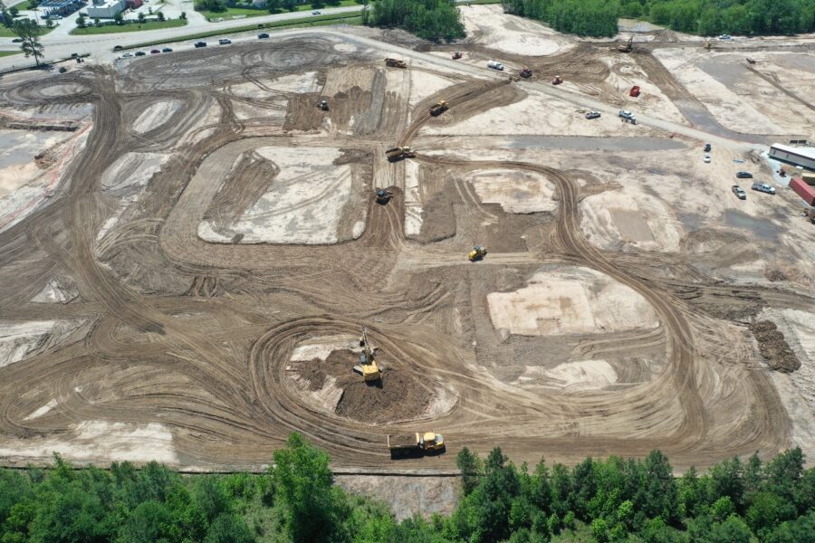 Aerial view of a large construction site with multiple excavators and bulldozers moving earth, creating dirt paths and cleared areas surrounded by trees and several parked vehicles.