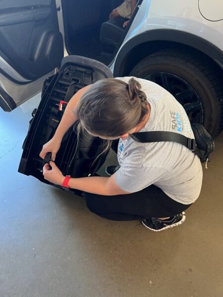 A person is kneeling on the ground next to a car, adjusting or inspecting a car seat with the car's rear door open.