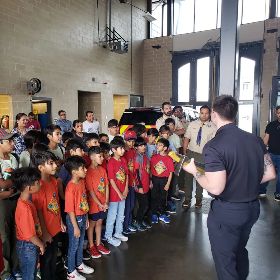 A group of children and adults listen to a man in uniform speak inside a garage-like building with large doors and a vehicle in the background.