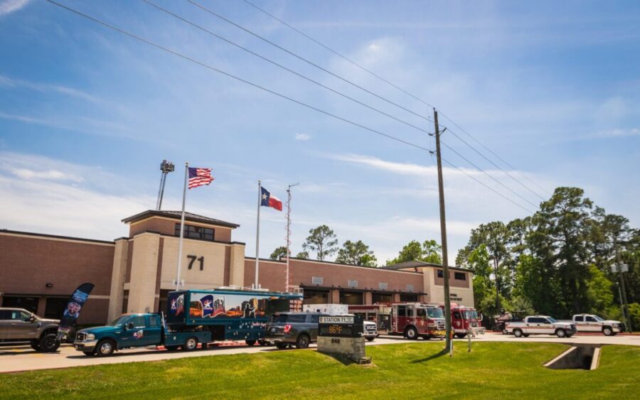 A fire station with the number 71 on the building, several emergency vehicles parked outside, and American and Texas flags flying.