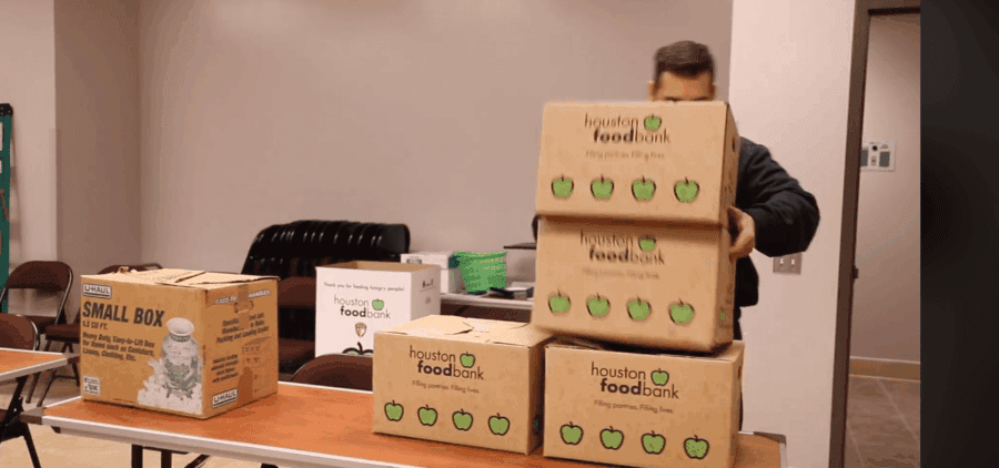 A person stacks boxes labeled "Houston Food Bank" and "small box" on a table in a room with chairs and other supplies.