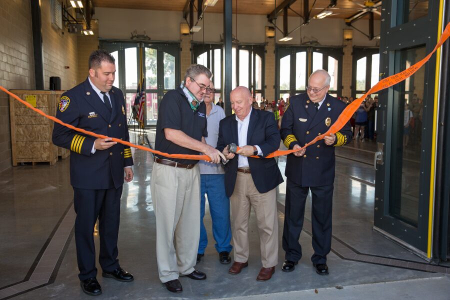 Four men, including two in dress uniforms, cut an orange ribbon at an indoor ceremony, with people visible in the background through open doors.