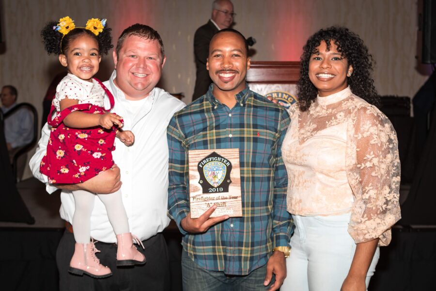 Four people pose together at an indoor event, with one man holding a plaque labeled "Firefighter 2019" and a woman holding a smiling child in a red dress.