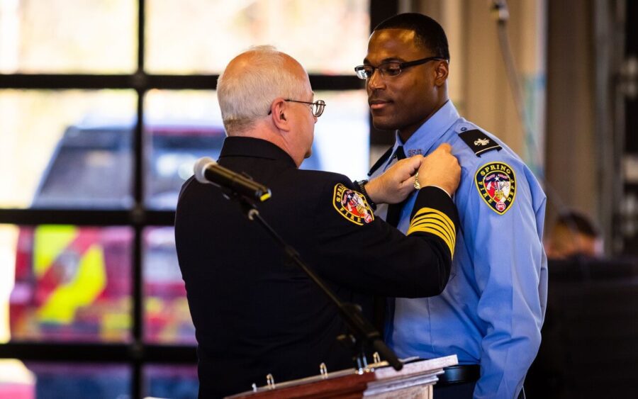 A uniformed officer attaches a pin to another officer’s shirt during a formal ceremony indoors, with emergency vehicles visible outside.