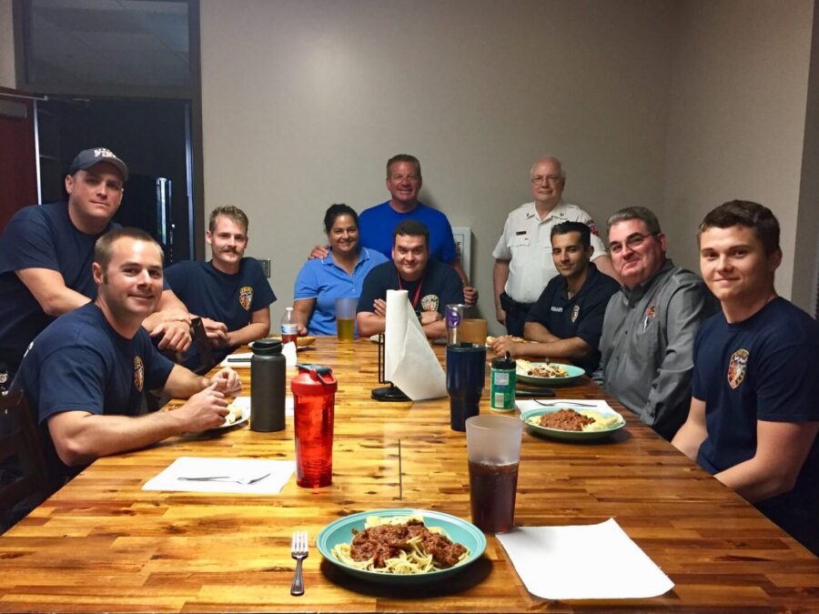 A group of people, including firefighters in uniform, sit around a table with plates of food, smiling at the camera in a dining room.