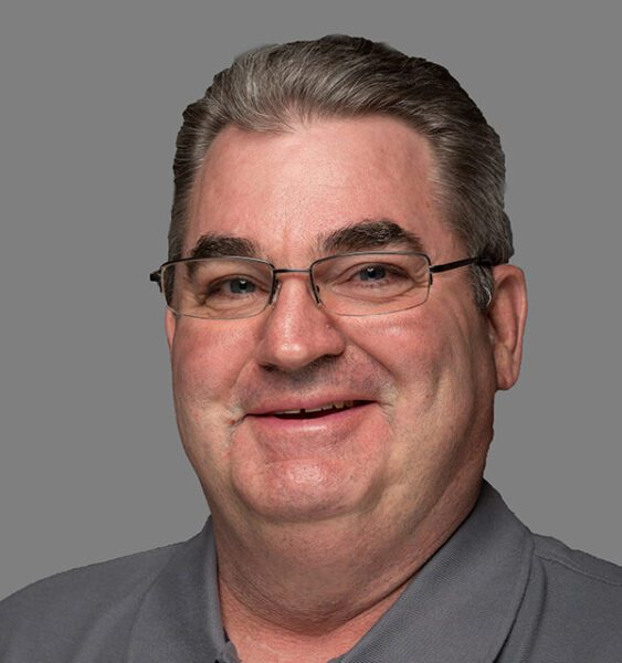 A middle-aged man with short gray hair, glasses, and a gray collared shirt smiles in front of a plain gray background.