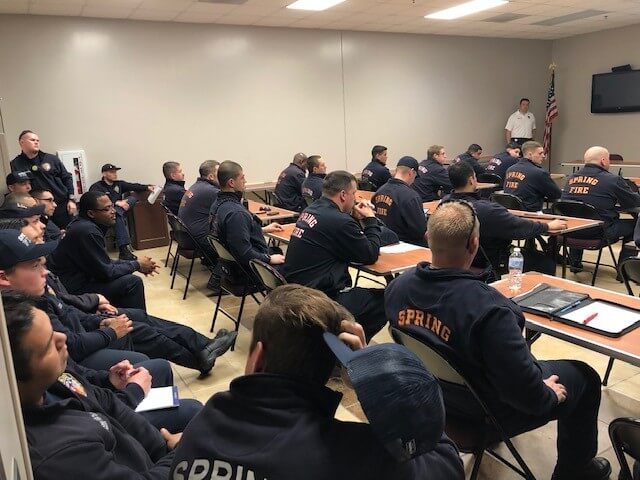 A group of firefighters in uniform sit in a classroom, listening to an instructor standing at the front near a screen and an American flag.