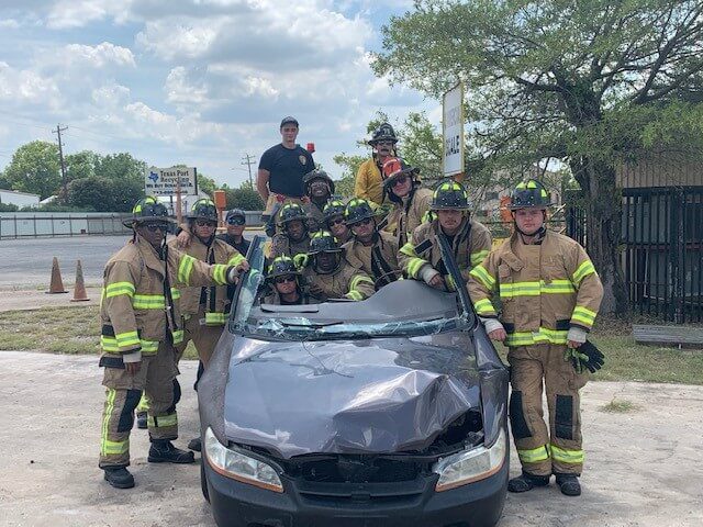 A group of firefighters in full gear pose around a damaged car with a removed roof, likely during a training exercise outdoors on a sunny day.