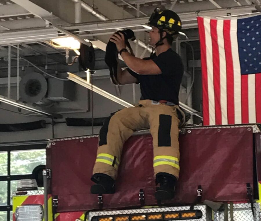 A firefighter in uniform sits on a fire truck, holding a small black cat. An American flag hangs in the background inside the fire station.